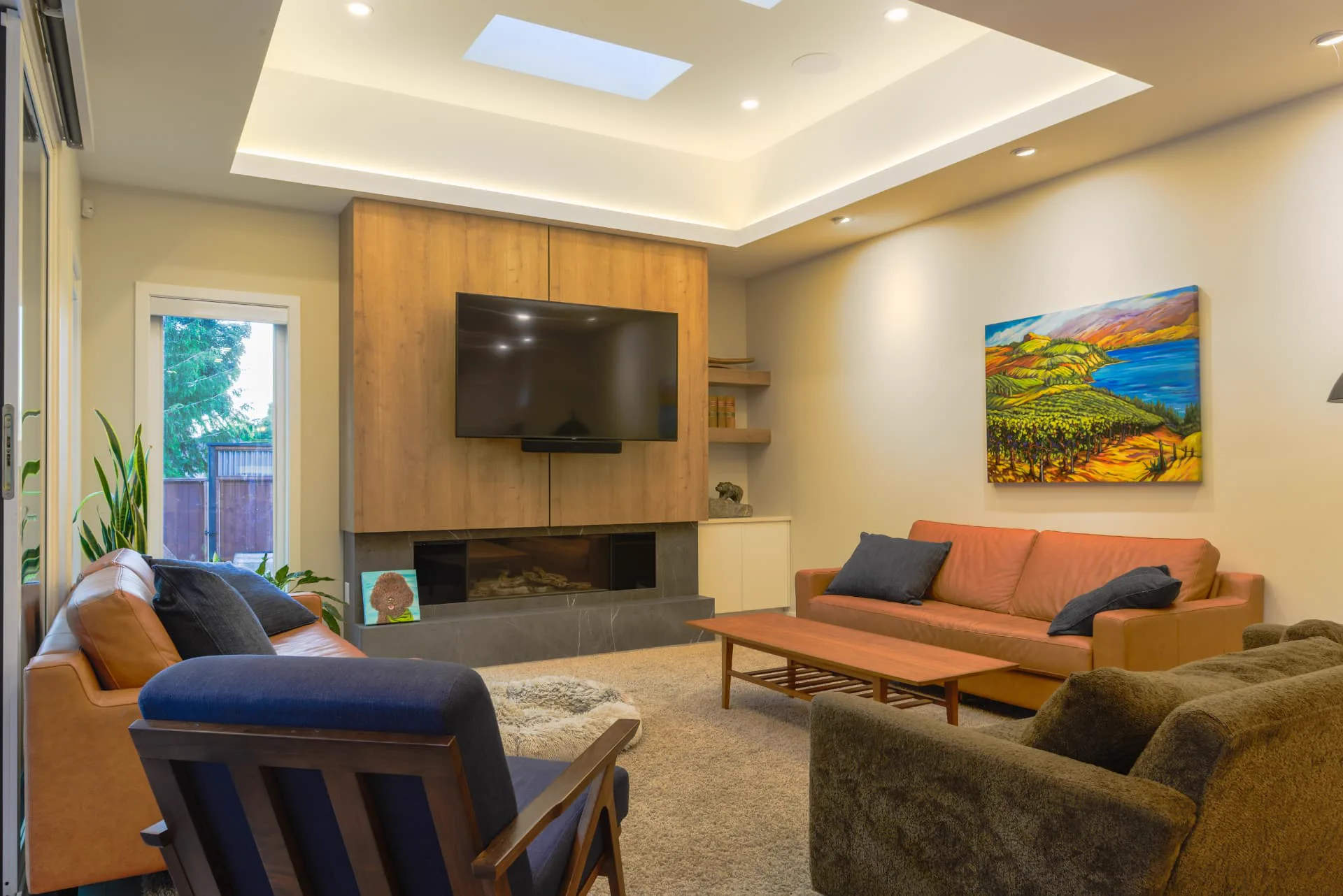Churchill family room, Penticton BC: leather sofas beneath a coffered ceiling with skylight, walnut media wall with linear fireplace and mounted TV, and a colourful Okanagan vineyard painting