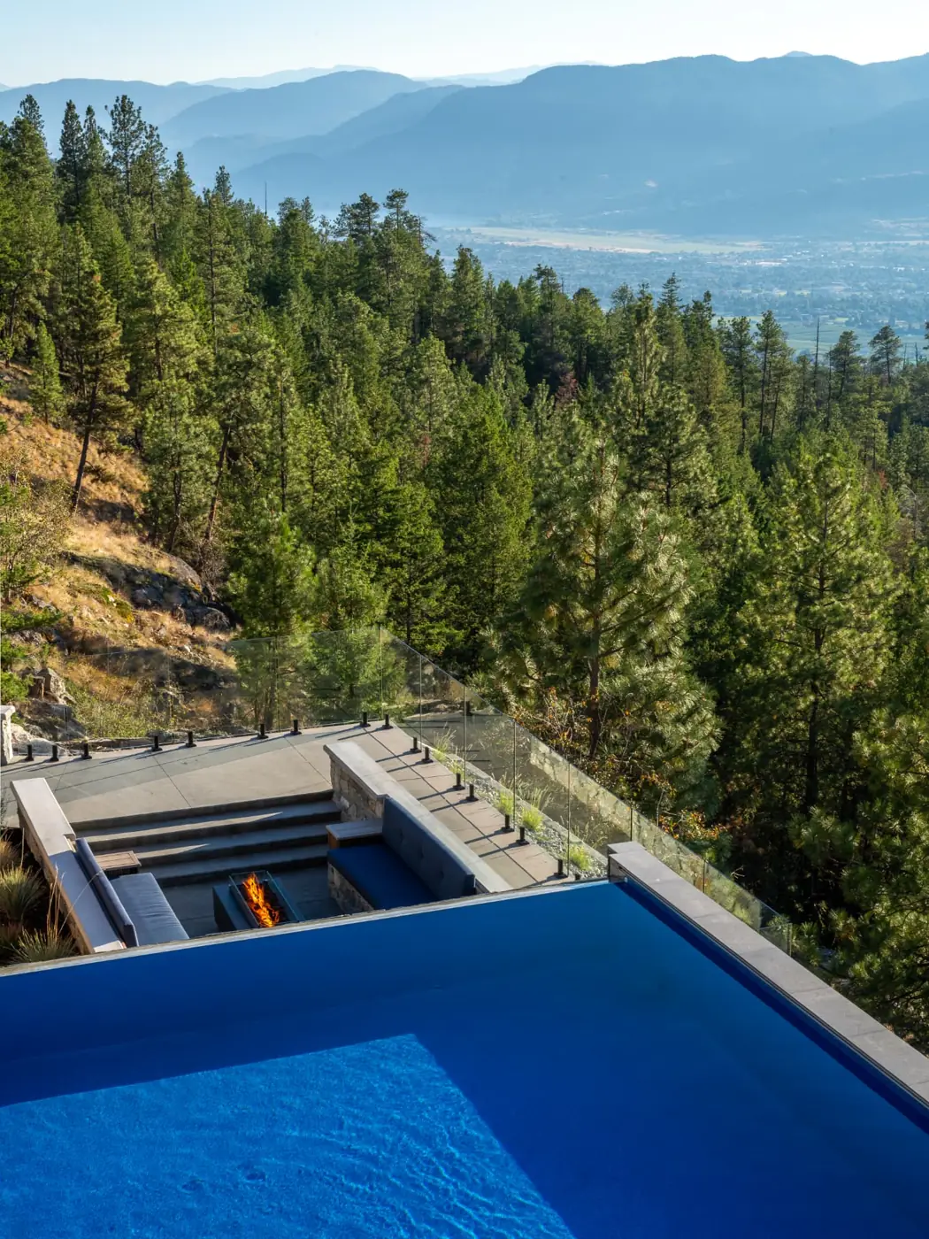 Vista infinity pool and sunken fire pit lounge with glass railing, Penticton BC, overlooking pine forest, the Okanagan valley and layered mountains