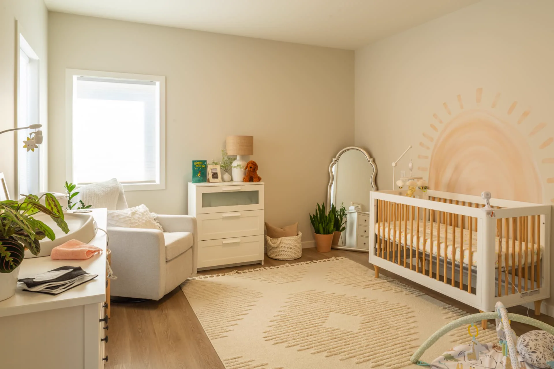 Ridge nursery, Penticton BC: sunlit nursery with oak-and-white crib, hand-painted sunrise mural, white dresser, and cushioned reading chair beside a garden window