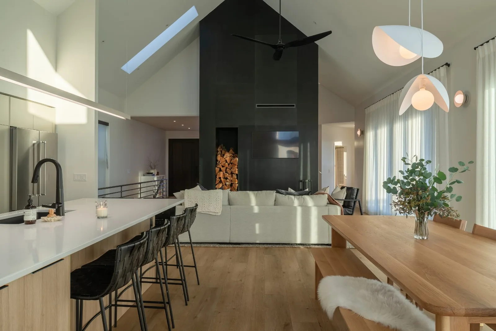 Abbott open kitchen and living area, Penticton BC: quartz island with black stools, blackened steel fireplace with firewood storage, and a vaulted ceiling with skylights