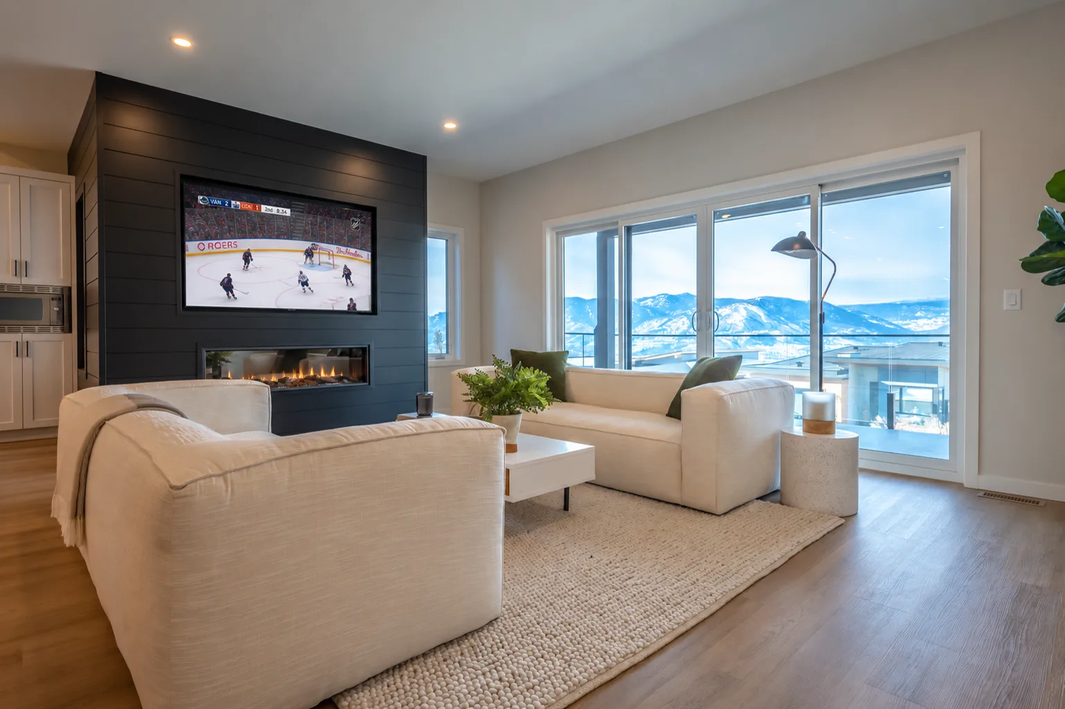 Fawn Court II living room, Penticton BC: cream sectionals on a wool rug, black shiplap media wall with fireplace and TV, and sliding glass doors framing Okanagan mountain views