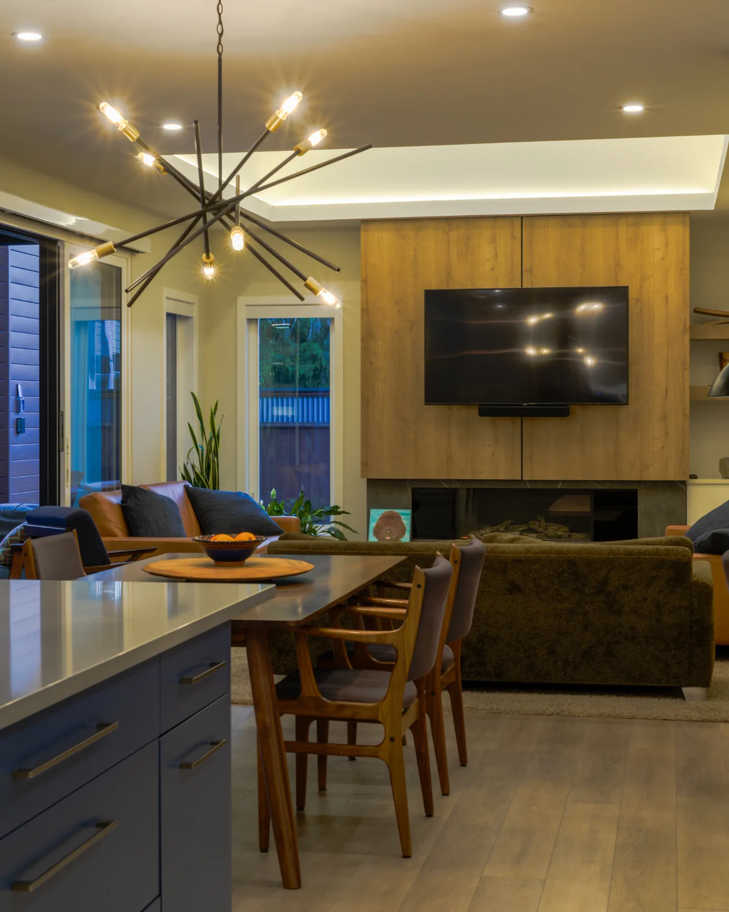 Churchill kitchen and dining room, Penticton BC: sputnik chandelier over a walnut dining table with mid-century chairs, blue cabinetry island, and olive sofa facing a wood-panelled media wall with fireplace
