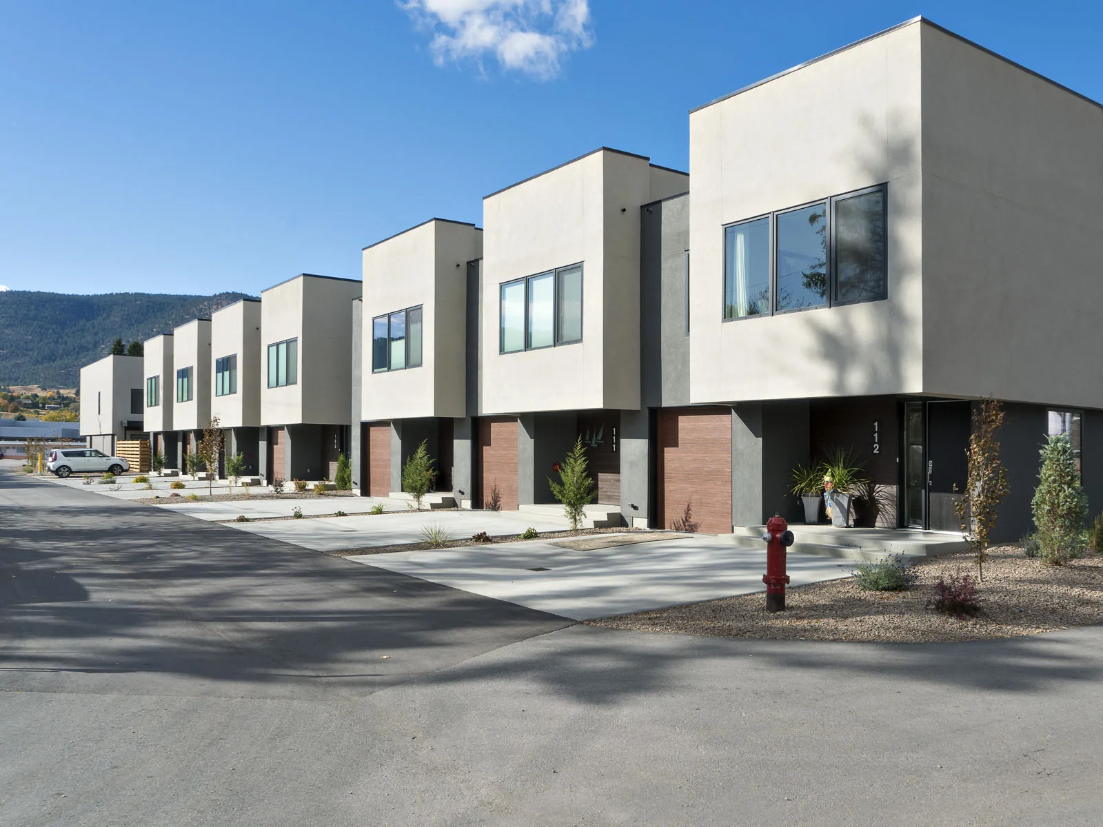 Beechwood townhomes exterior, Penticton BC: a row of modern stucco homes with wood-clad entries, individual driveways, and mountain views beyond