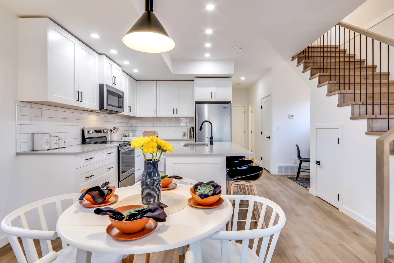 Hastings Ave kitchen and dining area, Penticton BC: white shaker cabinetry, subway tile backsplash, quartz island with black stools, and an open staircase