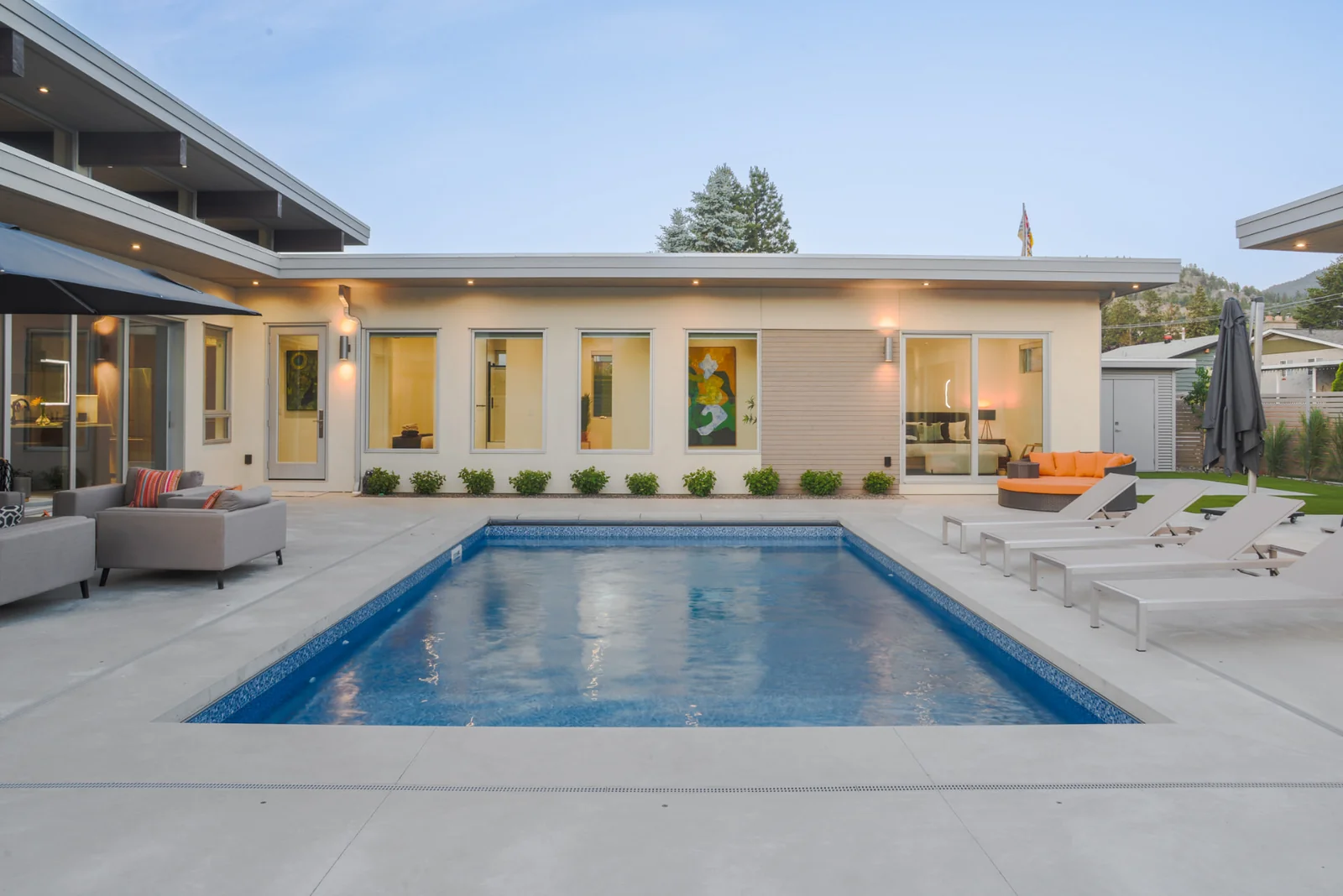Ridgedale backyard pool and exterior, Penticton BC: modern flat-roof home with carport, pool, patio seating, and landscaped lawn at twilight