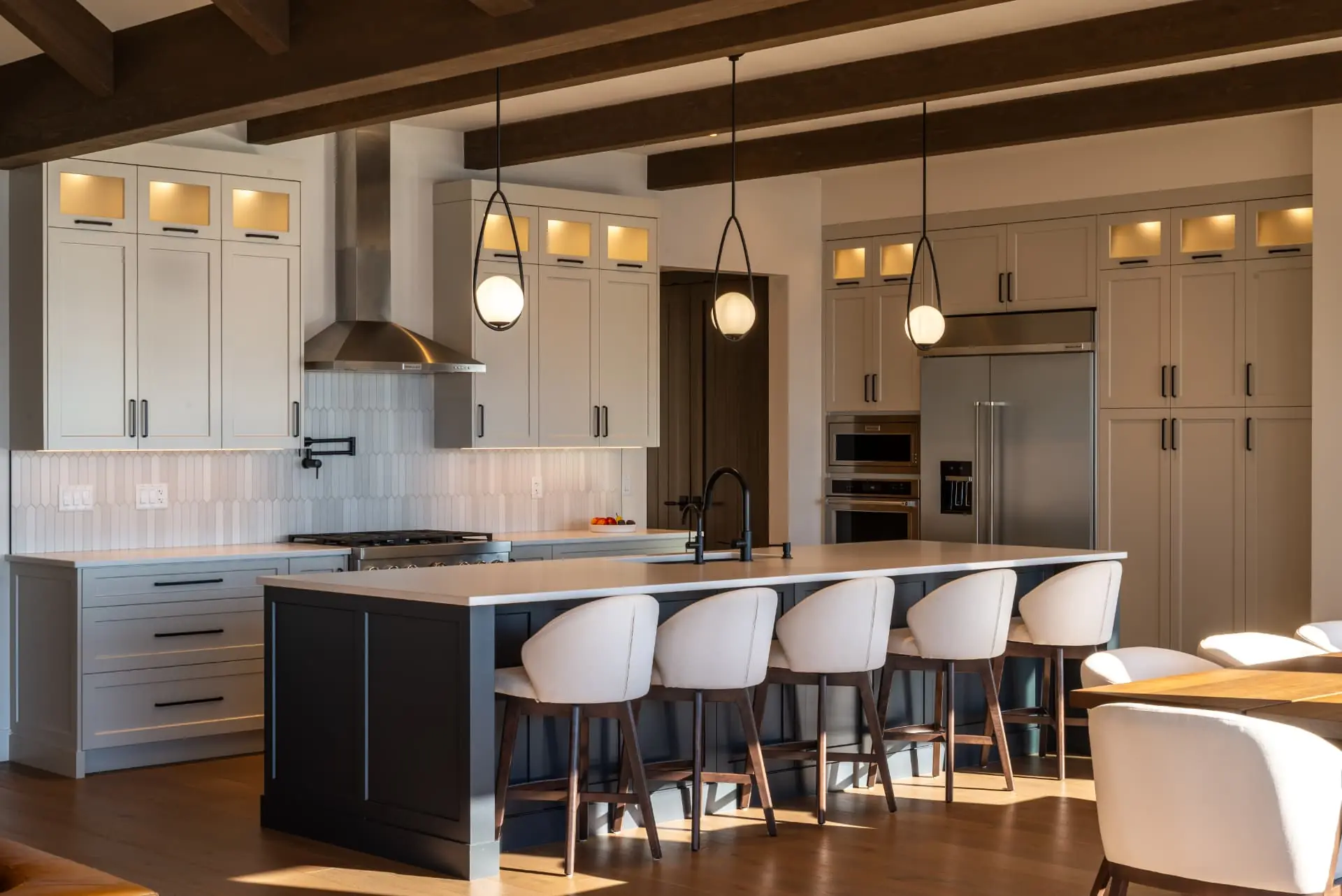 Vista kitchen, Penticton BC: long navy island with cream upholstered bar stools, white shaker cabinetry with lit glass uppers, globe pendant lights, stainless range hood, and dark timber ceiling beams