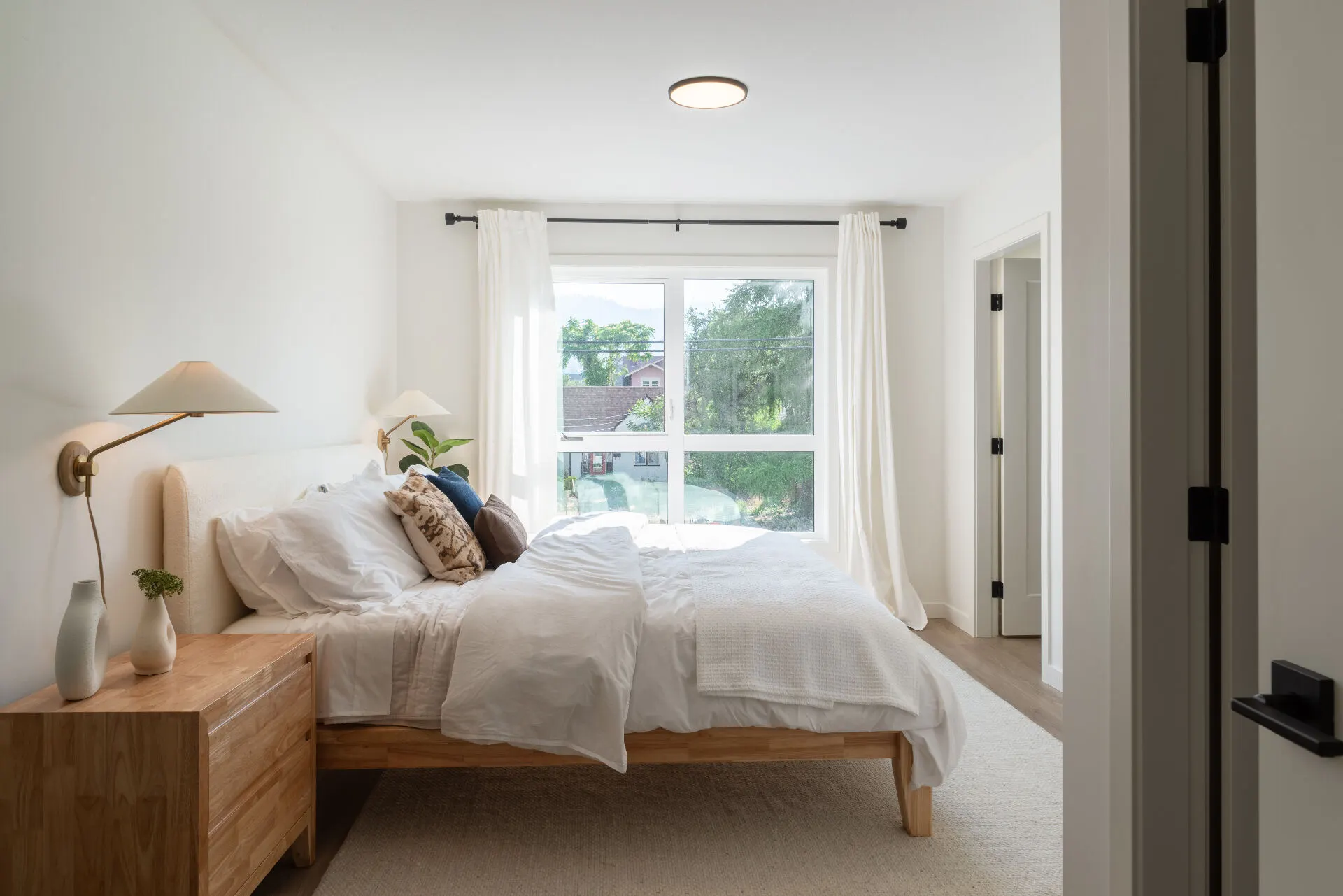 Urban Collection primary bedroom interior: oak bed frame, linen bedding, and a sunlit window framing the South Okanagan