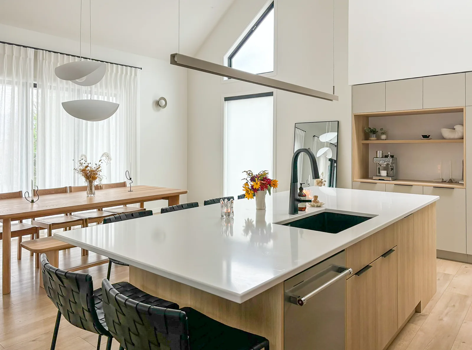 Abbott kitchen island, Penticton BC: white quartz counter, pale oak cabinetry, sculptural pendant lights, and an oak dining table by sheer-curtained windows