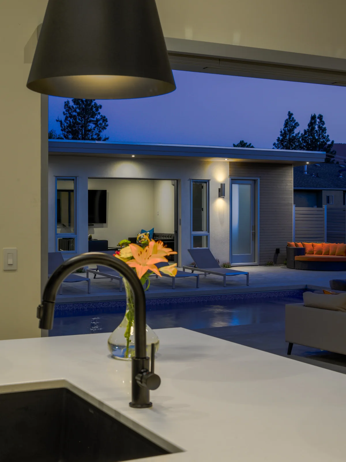 Ridgedale kitchen view, Penticton BC: matte black pendant and faucet over a white island, with lilies framing a twilight pool and modern flat-roof home beyond