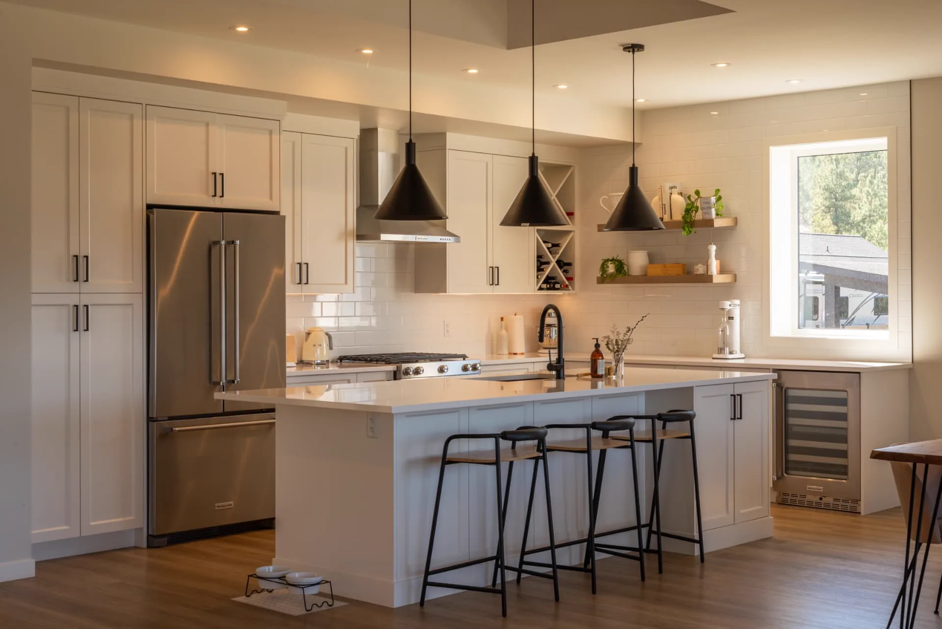 Ridge kitchen, Penticton BC: white shaker cabinetry, quartz island with black stools, matte black cone pendants, subway tile backsplash, and oak floating shelves