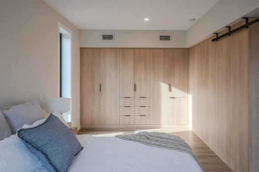 Basehome bedroom, Penticton BC: full-height oak closet wall, black barn door hardware, tall narrow window, and linen bedding in soft daylight
