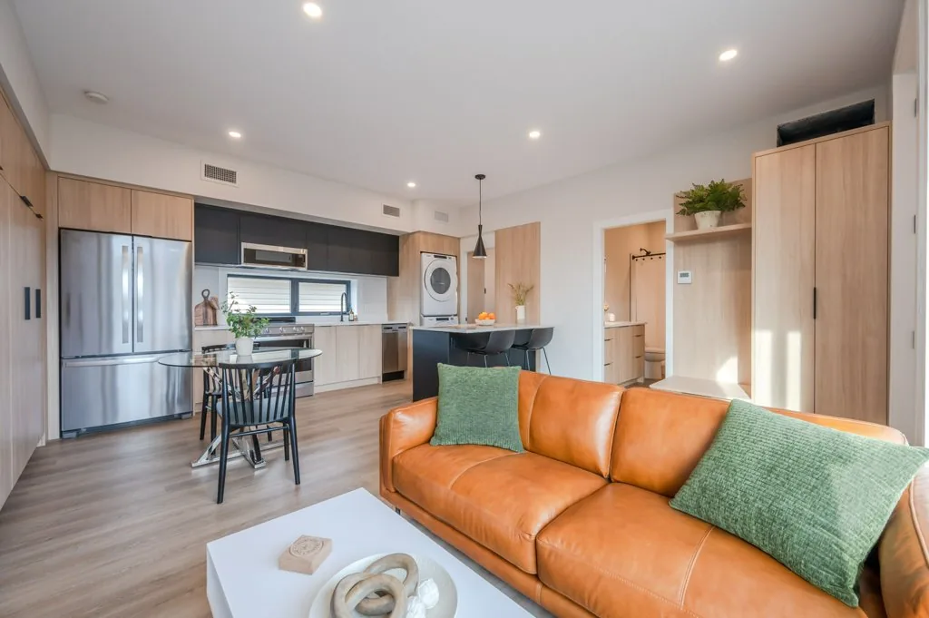 Basehome open kitchen and living room, Penticton BC: light oak cabinetry, stainless appliances, black island with bar stools, and a tan leather sofa on wide-plank floors