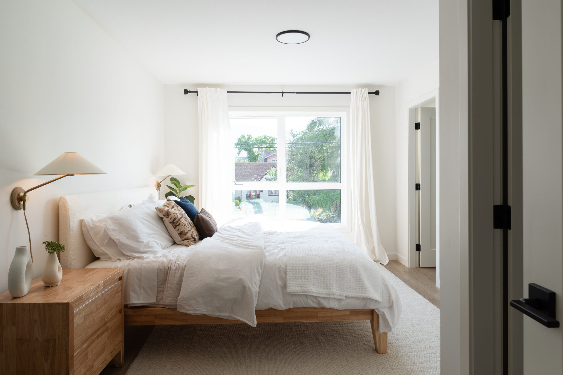 Light-filled Urban Collection primary bedroom with a boucle upholstered headboard, oak nightstand, brass sconces, and sheer linen curtains framing a sunlit Okanagan backyard.