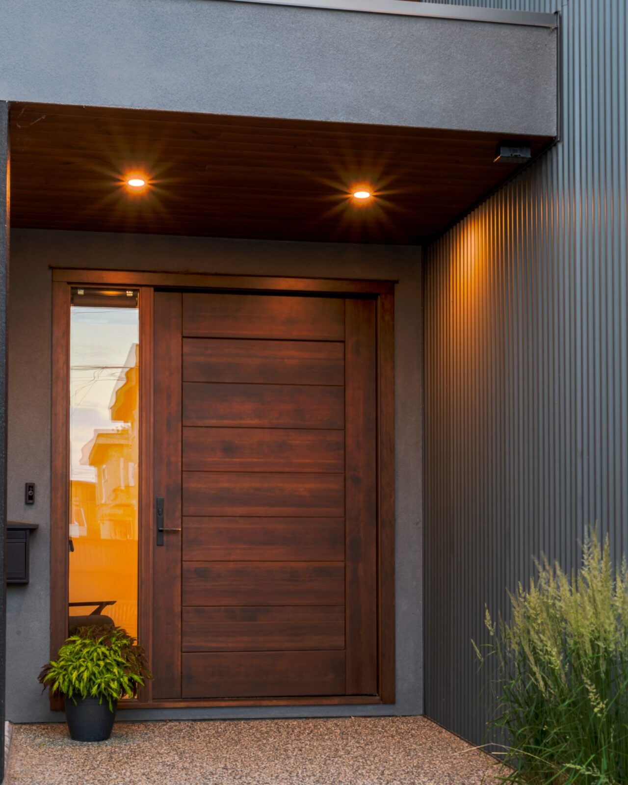 Custom Collection entry detail at the Churchill home in Penticton: warm walnut front door under warm downlights against charcoal stucco and corrugated metal siding by Parallel 50 Construction