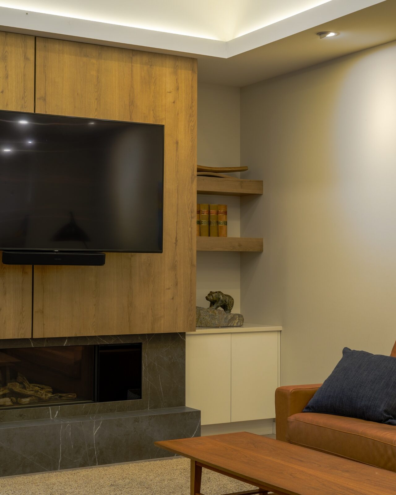 Custom Collection living room detail at the Churchill home: oak millwork media wall, honed marble fireplace surround, floating oak shelves, and a soapstone bear sculpture by Parallel 50 Construction